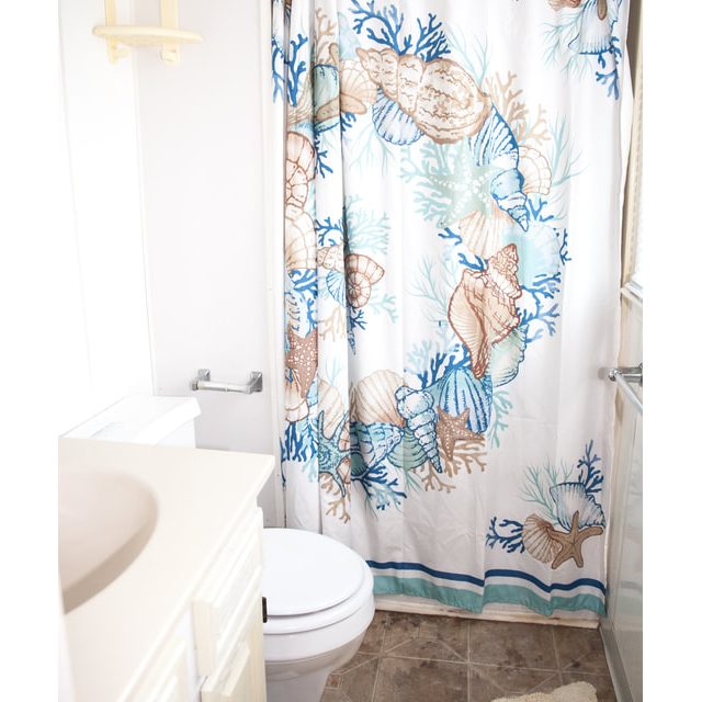 Small bathroom with seashell-patterned shower curtain, white toilet, beige vanity, and tan rug on tiled floor.