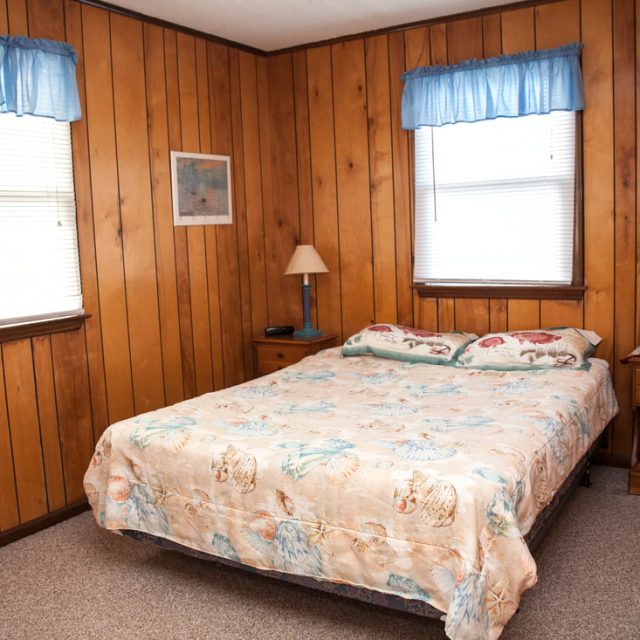 Small bedroom with wood-paneled walls, a double bed, two windows, a lamp, nightstand, chair, and books on a shelf.