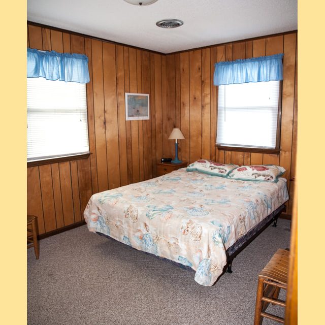 Small bedroom with wood-paneled walls, a bed with a seashell-patterned bedspread, two windows, and simple furnishings.
