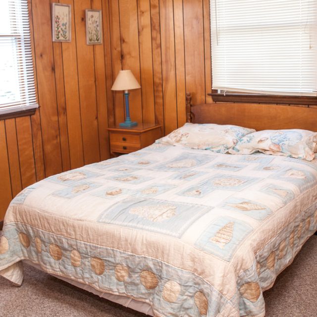 Small bedroom with wood-paneled walls, a double bed, light bedding, two windows, a nightstand, and a lamp.