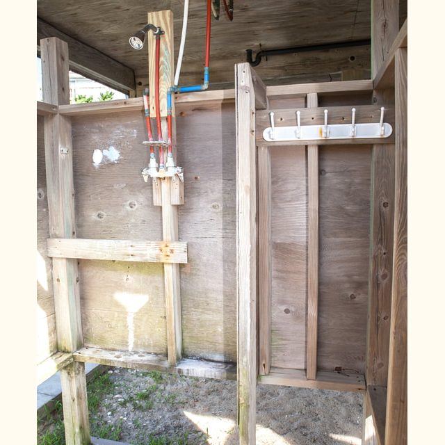 Outdoor wooden shower stall with exposed pipes, a showerhead, and a wall-mounted rack on the right side.