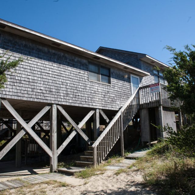 Gray wooden beach house on stilts with a staircase, surrounded by trees and sand under a clear blue sky.