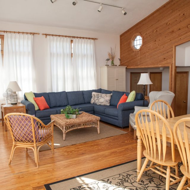 Living room with blue sectional sofa, wicker table, dining area, wooden floors, and sunlight through large windows.