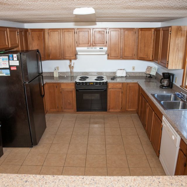 L-shaped kitchen with wooden cabinets, black fridge, stove, dishwasher, sink, coffee maker, and tile floor.