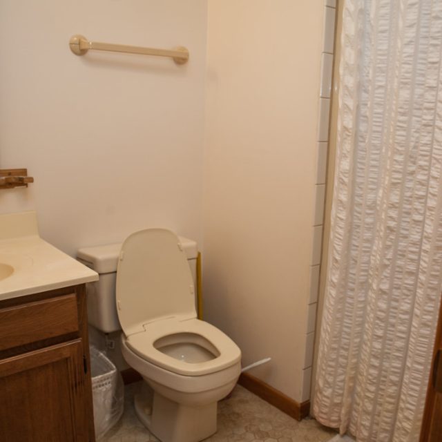 Small bathroom with a toilet, wooden vanity, wicker mirror, towel bar, and white shower curtain.