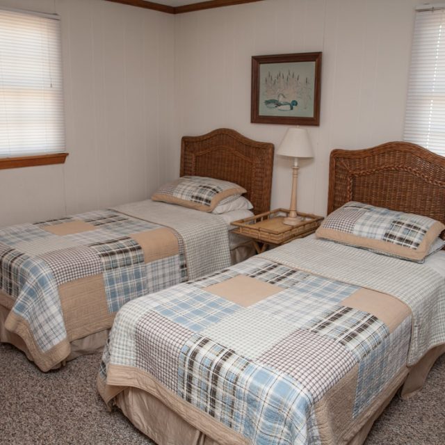 Small bedroom with two twin beds, plaid bedding, wicker headboards, nightstand, lamp, and three windows with blinds.