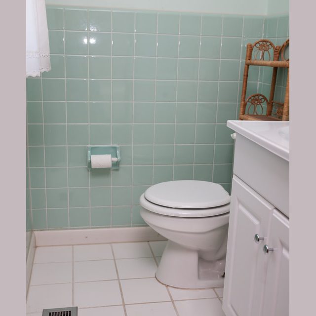 Bathroom with mint green tiled wall, white toilet, toilet paper holder, vent, and white vanity cabinet.