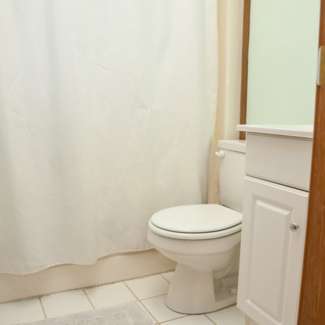 Small bathroom with white shower curtain, toilet, white vanity cabinet, and tile floor, viewed from an open door.