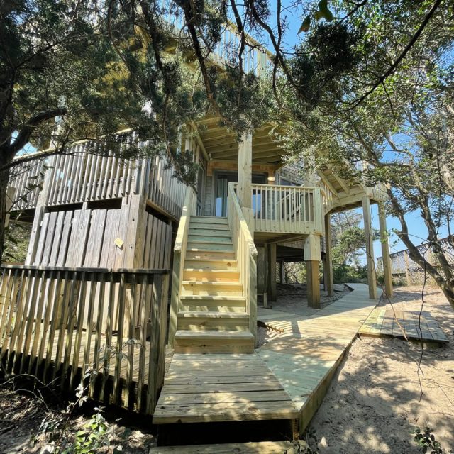 Two-story wooden treehouse with stairs and railings, surrounded by trees and sand under a clear blue sky.