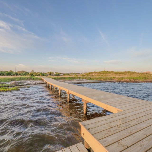 Wooden boardwalk extends over shallow water toward grassy shoreline under a clear blue sky with wispy clouds.