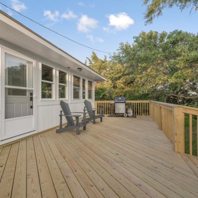 Wooden deck with two black chairs and a grill, attached to a white house, surrounded by trees under a blue sky.