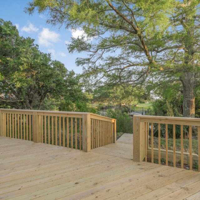 Wooden deck with railing overlooking green trees and a grassy yard on a clear, sunny day.