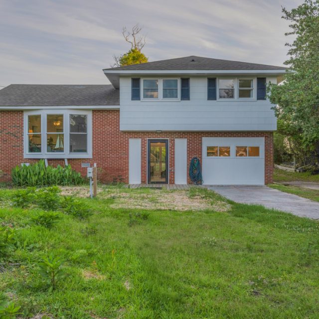 Two-story house with red brick and blue siding, front lawn, driveway, and garage door, surrounded by trees.
