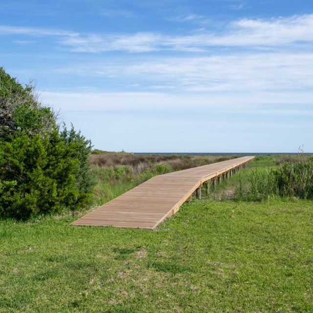 Wooden boardwalk stretching over grassy dunes toward the ocean under a blue sky with scattered clouds.