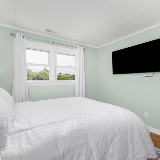 Minimalist bedroom with a white bed, light green walls, two windows, white curtains, and a wall-mounted TV.