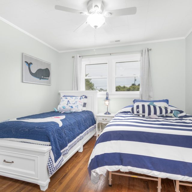 Two twin beds with nautical bedding in a bright bedroom with wood floors, white walls, and a ceiling fan.