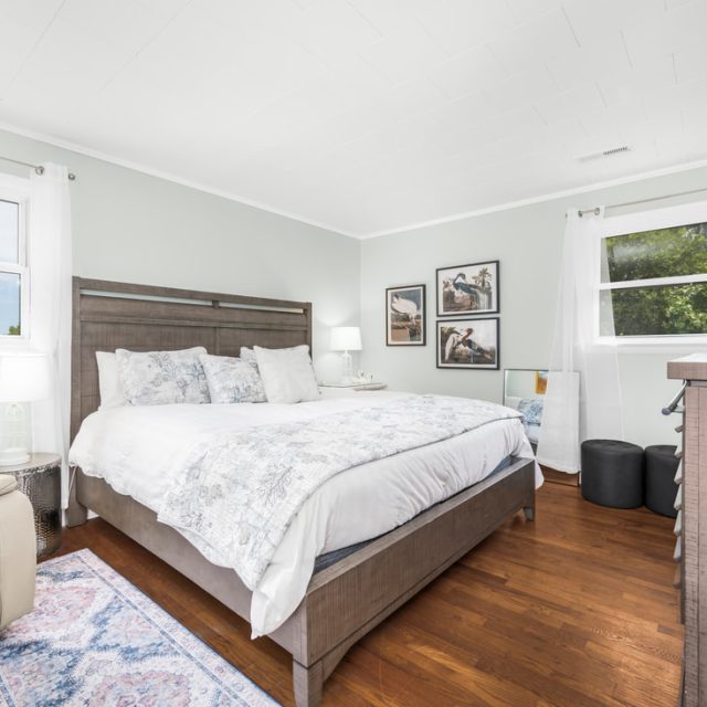 Bright bedroom with a large bed, wooden floor, armchair, dresser, wall art, and two windows showing trees outside.