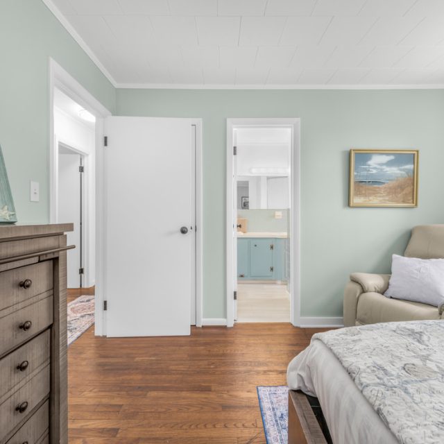 Bedroom with wooden floor, dresser, cream armchair, framed photo, bed, and doorways to a bathroom and hallway.