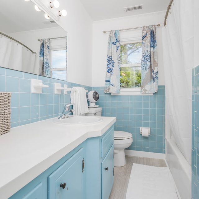 Bathroom with blue tile, white countertop, wicker basket, window with floral curtains, and a shower with white curtain.