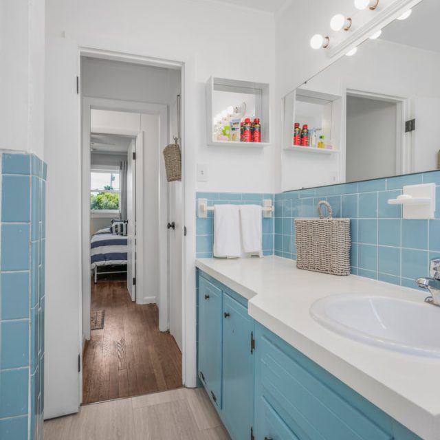 Bathroom with blue tile, white countertop, sink, mirror, and shelves; bedroom visible through doorway.