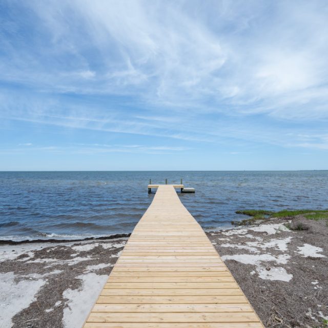 Wooden dock extending over calm blue water with partly cloudy sky and sandy shoreline in the foreground.