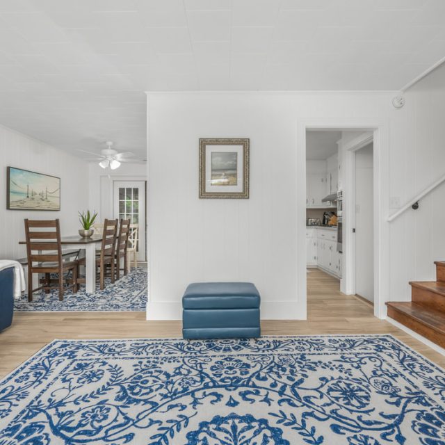 Bright living room with blue and white decor, wooden stairs, and a view into the dining area and kitchen.