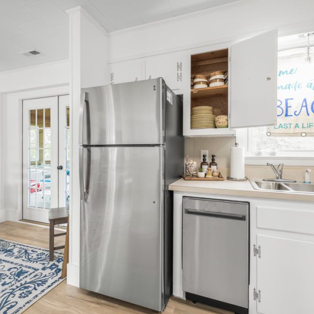 Modern kitchen with stainless steel appliances, open cabinet, and beach-themed decor near a dining area with wooden chairs.