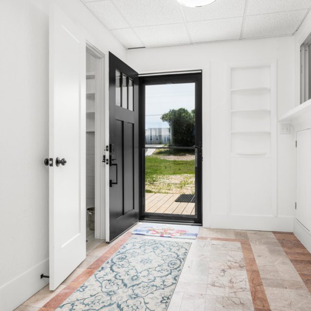 Bright entryway with open black front door, patterned rug, white walls, tiled floor, and built-in shelves.