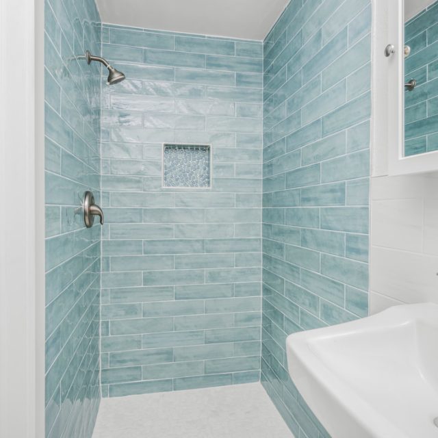 Walk-in shower with blue subway tiles, a built-in niche, and a white sink with a chrome faucet in the foreground.