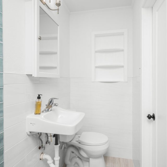 Small bathroom with a white sink, toilet, wall cabinet, shelves, and blue tile accent walls.