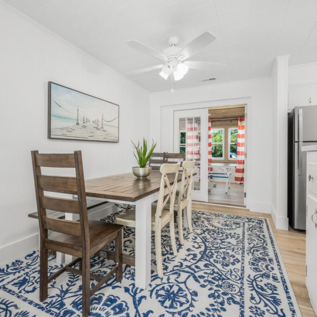 Dining area with wooden table, mixed chairs, blue patterned rug, and view into kitchen with stainless steel appliances.