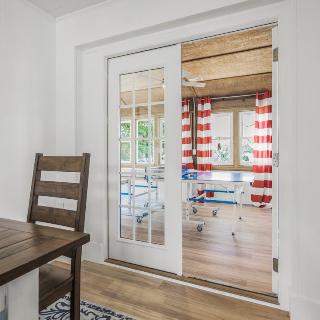 A glass door separates a small dining area from a sunroom with red and white striped curtains and wooden floors.