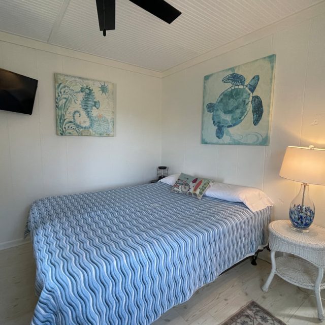 Bedroom with a large bed, blue patterned bedspread, wall art, lamp on a wicker nightstand, and a wall-mounted TV.