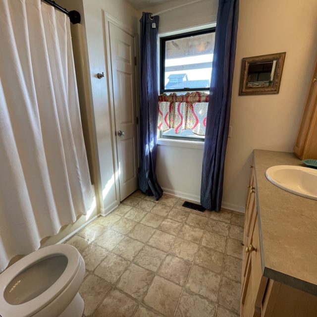 Bathroom with a toilet, sink, shower curtain, window with blue curtains, and beige tile floor.