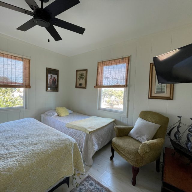 Small bedroom with two beds, armchair, wall-mounted TV, ship model, and three windows with bamboo shades.
