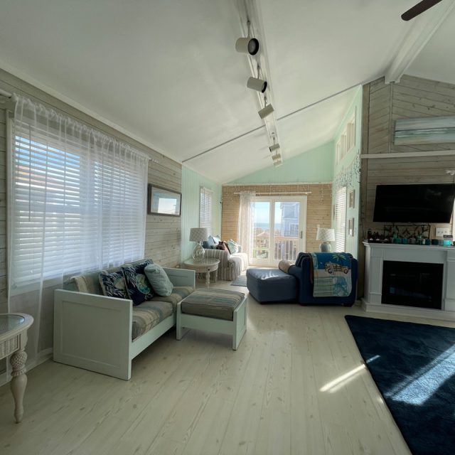 Bright living room with light wood floors, modern furniture, large windows, and a TV above a fireplace.