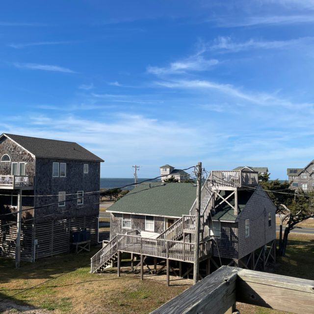 Wooden beach houses on stilts with decks, under a clear blue sky, near the ocean and power lines.
