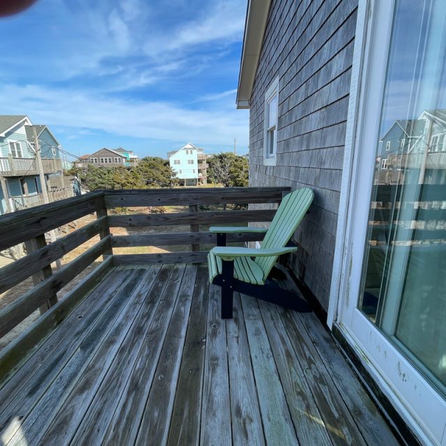 A green chair sits on a wooden deck outside a house, with neighboring houses visible under a blue sky.