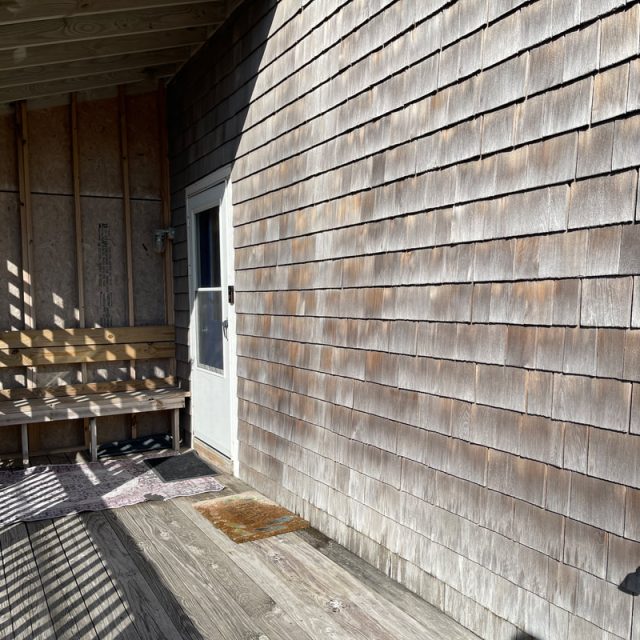 Wooden porch with a bench, chairs, rugs, and a door, all in sunlight with shadows from railings on the floor and wall.