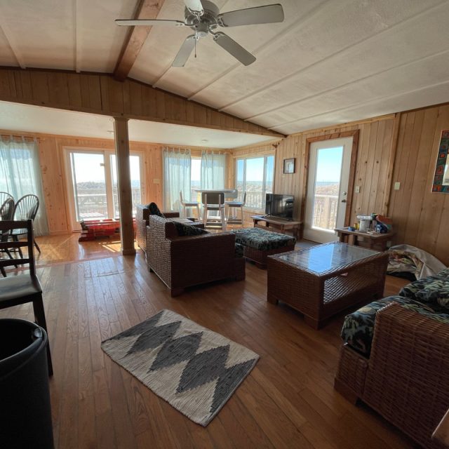 Living room with wood floors and walls, wicker furniture, large windows, and a ceiling fan. Dining area visible on left.
