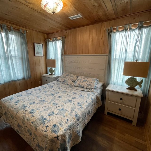 Wood-paneled bedroom with a bed, two nightstands, fish-shaped lamps, and sheer curtains on the windows.