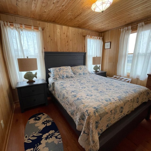 Wood-paneled bedroom with a double bed, two nightstands, lamps, and windows with white curtains.