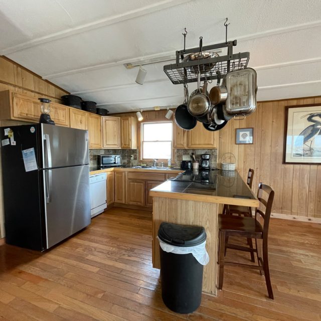 Wood-paneled kitchen with stainless steel fridge, dishwasher, hanging pots, and a small dining table with two chairs.
