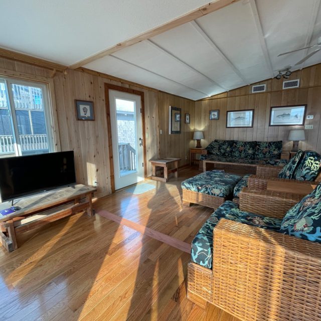 Sunlit living room with wood-paneled walls, wicker furniture, TV, and large windows showing an outdoor staircase.