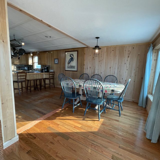 Dining area with a table and six chairs, wood-paneled walls, large window, and view into the kitchen.