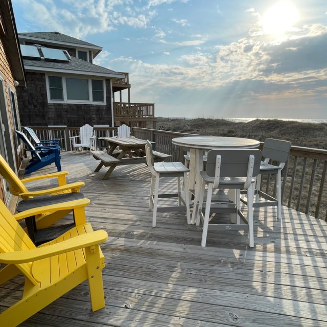 Wooden deck with yellow and white chairs, a table, and ocean view under a partly cloudy sky at sunrise or sunset.