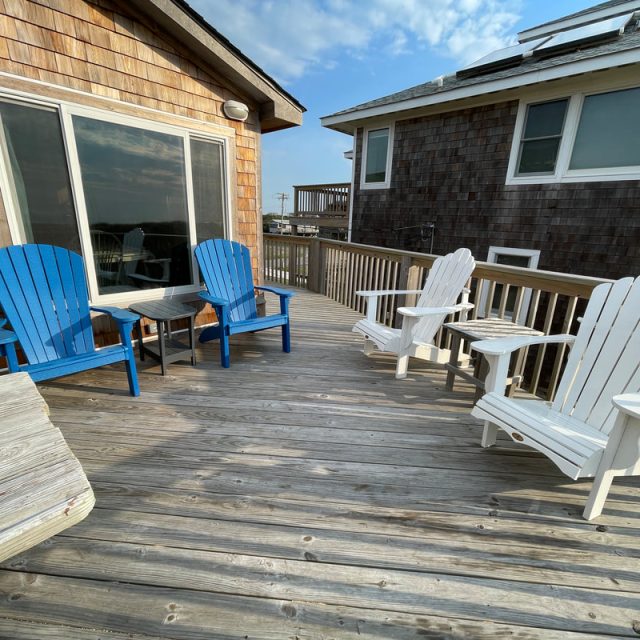 Wooden deck with two blue and two white Adirondack chairs, a small table, and two shingle-sided houses.