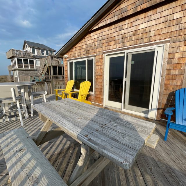 Wooden deck with a picnic table, colorful chairs, sliding doors, and a beach house in the background under a blue sky.