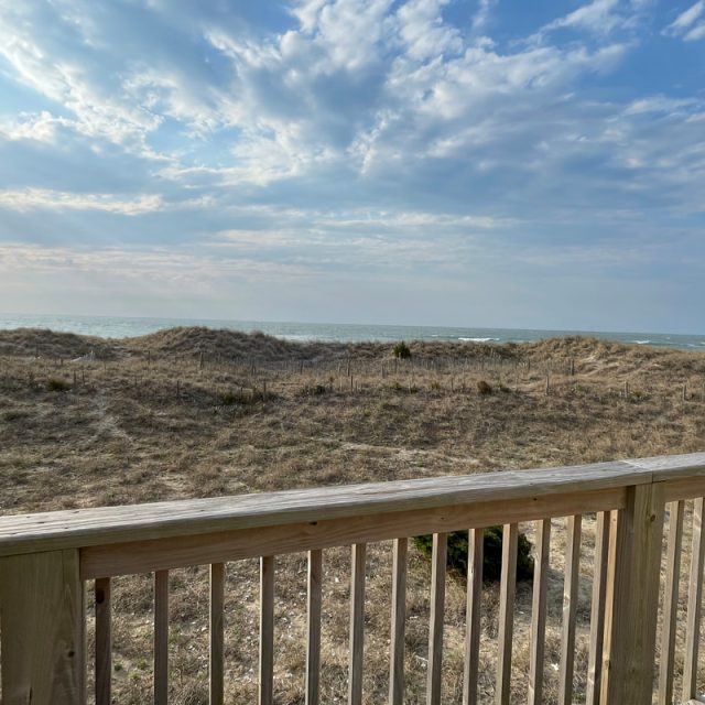 Wooden deck with railing overlooks sandy dunes, sparse grass, and ocean under a partly cloudy blue sky.