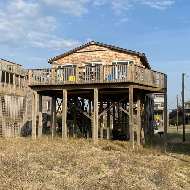Wooden beach house on stilts with a large deck, surrounded by dry grass and neighboring houses, under a partly cloudy sky.